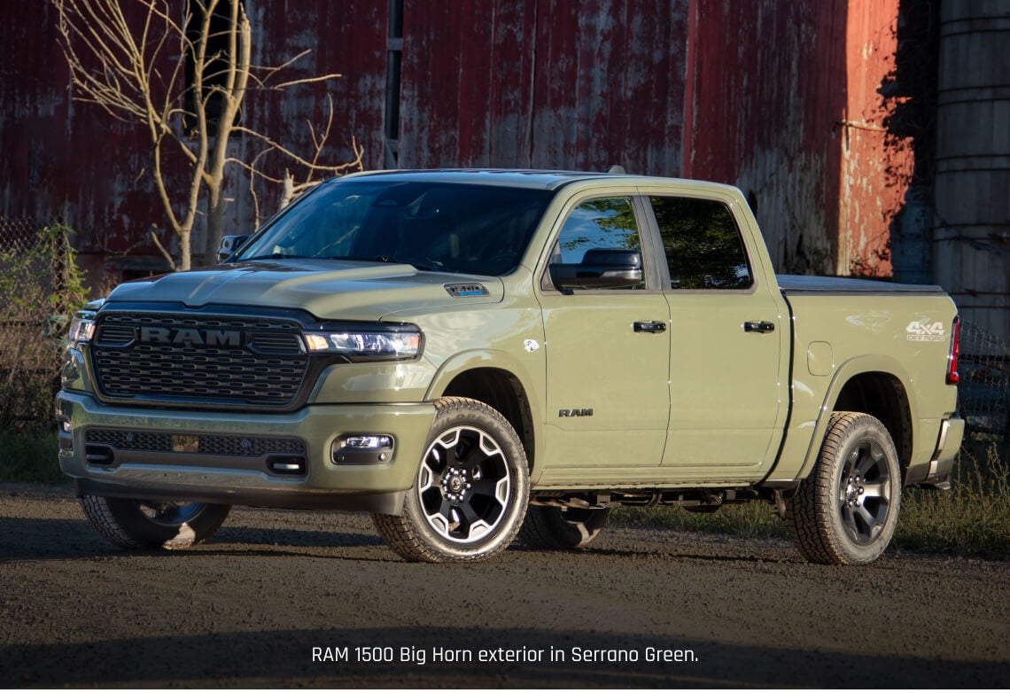 Front three-quarter view of a Ram 1500 Big Horn in Green parked on a dirt path by a rustic red barn.