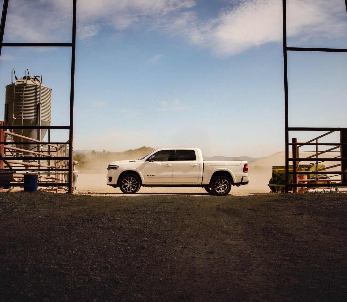 Side view of a white Ram 1500 framed by a barn doorway on a dusty farm field.