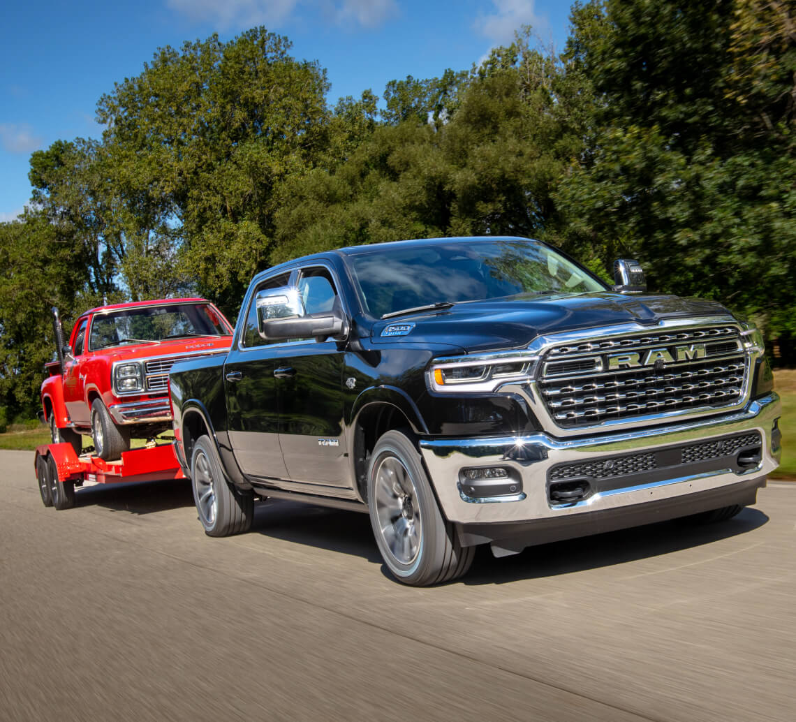 Front three-quarter view of a black Ram 1500 towing a red classic pickup on a paved road.
