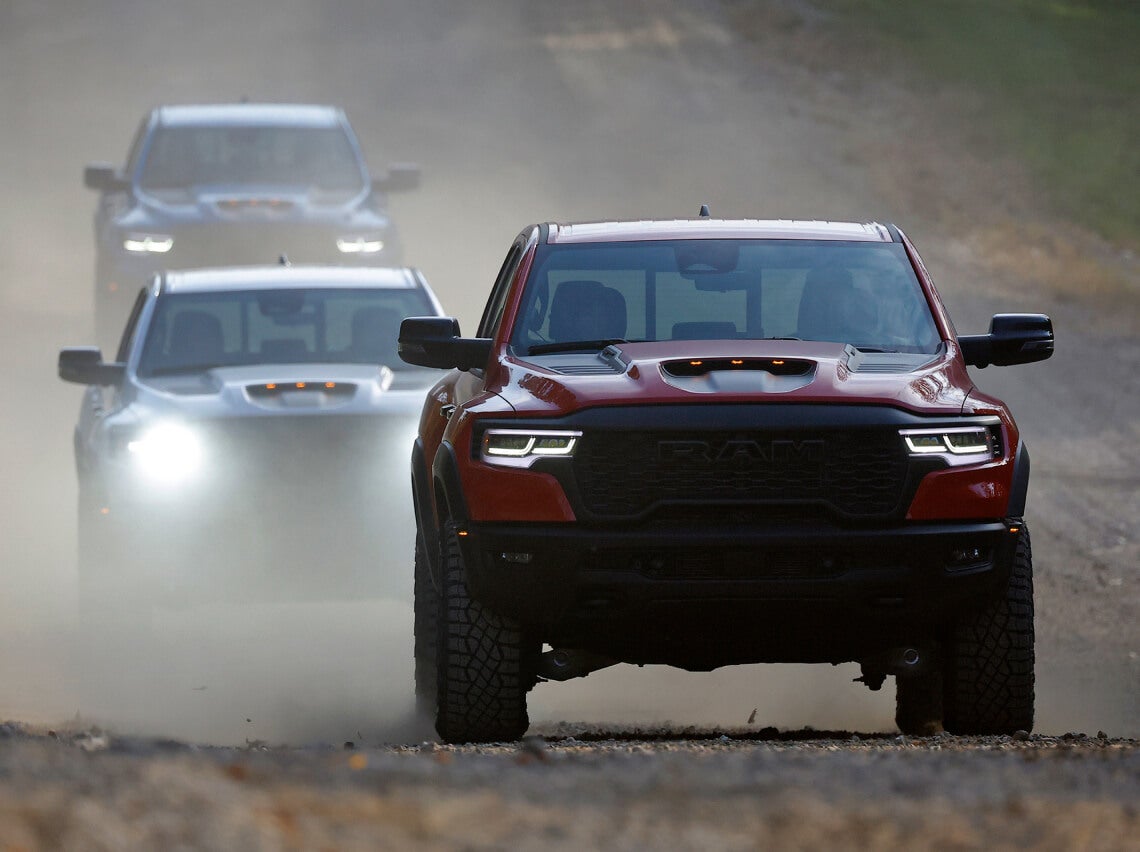 Red pickup truck kicking up dust on dirt road with vehicles in background